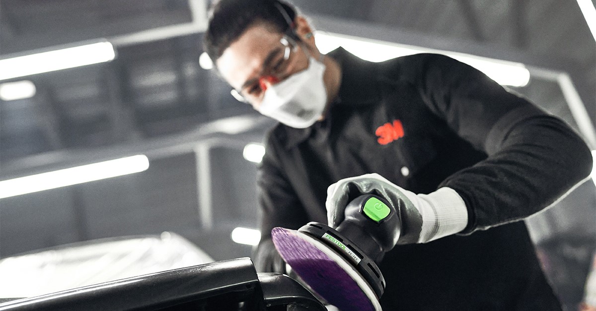 A man wearing safety glasses and a mask using an orbital sander to buff a car bumper.